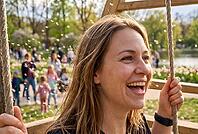 Swinging woman in public park, joyful recreation and mental wellbeing in urban green spaces, airborne dandelion seeds swirling in spring sunlight, carefree adulthood recapturing inner child freedom on wooden playground swings, lush lawn with strolling fam [IBR125414117]