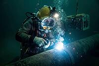 Subsea hyperbaric welding on industrial gas pipeline with commercial diving helmet and rising bubbles, offshore energy infrastructure maintenance, high risk deep sea repair combining specialized human craftsmanship with robotic remotely operated vehicle s [IBR125414118]