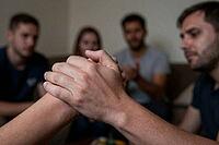 Interlocked hands forming a firm supportive grip represent collective solidarity and trust within a community, a blurred circle of companions observes the nonverbal agreement in an indoor environment, physical touch functions as a strong metaphor for ment [IBR125414122]