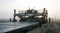 Heavy concrete paving machine expanding highway infrastructure, civil engineering workers supervising road construction on industrial site at sunrise, massive slipform paver laying smooth concrete slab across wide transportation route, construction crew w [IBR125414126]