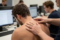 Ergonomic spine examination and occupational health physiotherapy for desk worker suffering back pain and cervical strain in digital workspace, hands of therapist gently palpating trapezius muscles and upper cervical vertebrae of shirtless young employee  [IBR125414128]