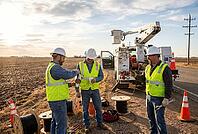 Utility linemen take a coffee break beside cable spools and a cherry picker truck during rural infrastructure expansion and power grid maintenance, technicians in high visibility gear collaborate on telecommunication line installation along open farmland, [IBR125414137]