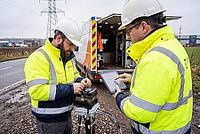 Fiber optic broadband deployment, telecommunication technicians install digital network infrastructure beside roadside utility van, team engineering, field technicians wearing high visibility safety gear inspect fiber cables using fusion splicer and table [IBR125414159]