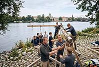 Collaborative timber construction and intergenerational craftsmanship on Potsdam riverbank, youth education outdoor workshop experiential learning, knotting ropes and stabilizing wooden beams with teamwork, hands on pedagogy fostering manual skills across [IBR125414168]