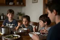 Family mealtime with siblings eating warm lunch together around rustic dining table using wooden chopsticks, nourishing home cooked food in bowls, quiet domestic gathering during daytime, child nutrition, commensality as social cohesion in everyday home l [IBR125414178]