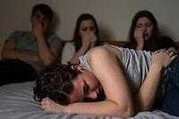 Mental health crisis, emotional trauma, depression, grief among youth, young adult crying on bed while friends react in shock and disbelief, overwhelming psychological distress within a close group of peers, somber atmosphere of vulnerability and sudden l [IBR125414179]