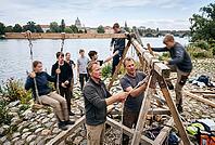 Experiential outdoor education and manual teamwork by the river, youth group and mentors constructing a wooden swing structure with ropes and timber, intergenerational knowledge transfer and vocational craft skills in social pedagogy, active collaboration [IBR125414182]