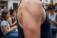 Athletic male shoulder and muscular arm in sleeveless shirt standing in crowded urban street setting during warm summer season, skin texture and anatomical definition highlighting body culture and physical wellness in contrast to digital distraction, blur [IBR125414204]