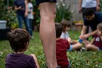 Intergenerational community gathering on green garden lawn with adult leg standing as protective pillar over playful children learning social skills, domestic childcare and pedagogical development in familial microenvironments, parental supervision repres [IBR125414210]
