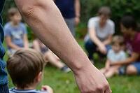 Protective adult guidance overlooking children playing on green grass in social community gathering, intergenerational childcare and outdoor education, a prominent masculine forearm in the foreground frames blurred family members and toddlers seated toget [IBR125414211]