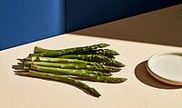Fresh green asparagus spears rest on a beige surface. Blue wall behind adds contrast to composition. Small white plate nearby for serving or styling, AI generated [IBR125421254]