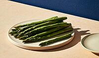 Fresh green asparagus spears on white ceramic plate. Plate rests on light beige surface. Deep blue wall behind adds contrast, AI generated [IBR125421261]