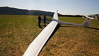 Two people standing near a glider on a grassy field with mountains in the distance on a sunny day, Aérodrome de Château-Arnoux - Saint-Auban, France [IBR125421624]