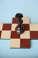 A dark wooden queen stands on a brown and beige chessboard against a light blue background [IBR125424489]