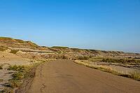 An asphalt road curves through a vast landscape of eroded hills and layered rock formations under a bright, cloudless sky, Drumheller, AB T0J 0Y0, Alberta, Canada [IBR125424505]