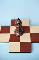 A wooden brown bishop on a brown and beige chessboard against a light blue background [IBR125424512]
