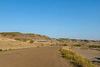 A paved road curves through a vast landscape of eroded hills displaying distinct geological strata under a bright, cloudless sky, Drumheller, AB T0J 0Y0, Alberta, Canada [IBR125424515]