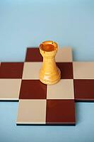 A yellow wooden rook stands on a brown and beige chessboard against a light blue background [IBR125424535]