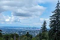A vast urban landscape stretches towards the horizon under a dramatic sky filled with clouds, framed by lush green conifers, Centennial Way, Burnaby, BC, Canada [IBR125424548]