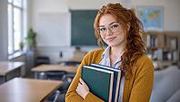 A smiling redheaded student with freckles and clear glasses holds books in a sunlit classroom, wearing a mustard cardigan over a striped shirt, AI generated [IBR125424575]