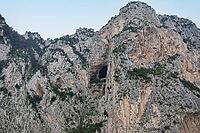 Rocky mountain cliff face displaying a prominent cave opening against a clear sky [IBR125436240]
