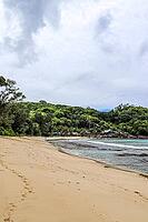 Sandy beach on tropical island showing turquoise ocean water, green jungle, and hillside architecture under cloudy sky [IBR125436258]