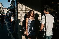 Geometric grid shadows fall upon a contemplative young woman during an emotional conversation with a man on a bustling European city street in summer, architectural latticework projections cast a psychological cage across her face evoking themes of emotio [IBR125446413]