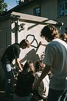 Youth subculture and communal connection thrive outdoors, young adults gathering near urban walls with surveillance cameras and graffiti artwork, candid camaraderie merges with societal tension beside a utility pavilion, harsh diagonal afternoon sunlight  [IBR125446419]