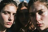 Three young women gazing intensely into lens under harsh sunlight, strong facial shadows create an intimate study of identity, vulnerability and modern youth solidarity, the central figure recedes into deep shade between two brightly illuminated faces fra [IBR125446422]