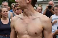 Athletic shirtless young man stands amidst a dense public crowd outdoors during summer, displaying physical presence, vulnerability, exposed muscular torso, attentive onlookers observing the gathering, collective gaze focusing on individual strength, anat [IBR125446430]
