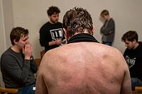 Vulnerable shirtless man surrounded by supportive peers in emotional group therapy session experiencing anxiety tension vulnerability and catharsis, raw human exposure manifests through physical posture and bare shoulders confronting unresolved collective [IBR125446436]