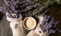 Cup of coffee sits surrounded by dried lavender sprigs. Lavender flowers rest on a dark wooden surface. White tissue paper wraps some lavender bundles, AI generated [IBR125446439]