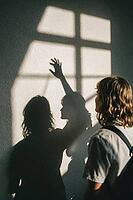 Silhouetted hand reaching toward window light cast on concrete wall, intimate human connection, longing, shadow play, emotional duality, introspective youth, artistic framing through natural sunlight, contrasting contours of two figures projecting their i [IBR125446441]