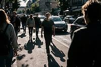 Urban alienation and commuter solitude characterize modern city life as busy sidewalks merge with congested vehicle traffic during late afternoon, a contemplative male pedestrian strides forward along the sunlit asphalt while stark elongated silhouettes p [IBR125446442]