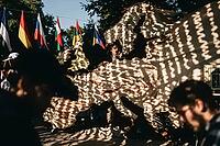 Performer behind a large cutout textile map of Europe during an outdoor theatrical event, dynamic dappled sunlight creating intricate shadow patterns across continental landmasses, vibrant international national flags suspended along a tension wire in the [IBR125446443]