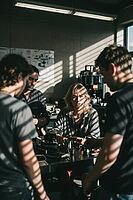 Focused young artisans collaborate around a metallic workbench analyzing precision equipment under dramatic Venetian blind shadows, an attentive blonde woman sits centrally immersed in critical evaluation while three colleagues lean forward in shared conc [IBR125446448]