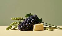 Dark purple grapes sit beside a wedge of hard cheese. Green wheat stalks rest nearby. Soft light highlights textures, AI generated [IBR125446450]