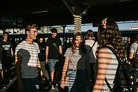 Commuters traverse railway platform during rush hour, rhythmic slatted shadows cast across young travelers standing in contemplation, transit passengers experiencing quiet anticipation within public transport networks, warm sunlight filtering through stat [IBR125446454]
