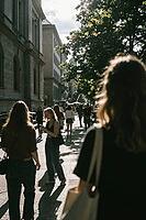 Pedestrians navigate busy urban sidewalks bathed in intense summer sunlight and contrasting shadow, mobile phone distraction reflects modern digital isolation, historical stone architecture flanks the crowded thoroughfare while leafy canopy foliage filter [IBR125446456]