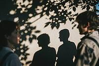 Interpersonal resonance and emotional projection, young people standing opposite each other while their dark silhouettes cast an intimate dialogue upon a bright stucco facade, dappled foliage contours intertwining with human profiles, subconscious nonverb [IBR125446457]