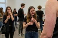 Authentic vocal training and performing arts education in a minimalist studio, young adults engaging in emotional communication exercises, expressive body language and speech practice, an expressive young woman gesturing nervously near her chest during an [IBR125446458]