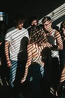 Subtle emotional tension, vulnerability and group cohesion unfold among young adults gathered in dramatic natural window light through blinds, casting parallel shadow stripes across bodies and bare skin, introspective glances and contemplative facial expr [IBR125446464]