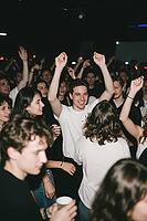 Ecstatic youth celebrating togetherness on a crowded nightclub dancefloor, joyful party culture embodying social cohesion and vitality, a smiling young man in a white shirt raises both arms into the air as a profound expression of liberation, uninhibited  [IBR125446466]