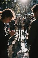 Digital connectivity meets urban isolation amidst heavy rainfall, pedestrians fixate on glowing smartphones while traversing wet city pavement, backlighting illuminates falling raindrops and casts long cast shadows along drenched asphalt, young men in the [IBR125446467]