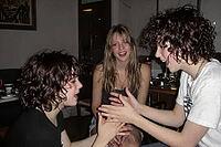 Three young friends enjoying playful hand games during an evening gathering, sharing authentic joy, laughter and emotional connection, dynamic gesture coordination between two companions clapping hands while a smiling girl observes with cheerful amusement [IBR125446471]