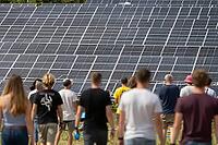 Civic participation in renewable energy transition, people walk toward massive photovoltaic solar panels across open grassland during bright daylight, collective human momentum aligns with industrial clean power infrastructure to symbolize ecological awar [IBR125446474]