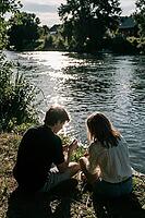 Youthful romance, tranquil riverbank intimacy, sparkling water reflections of summer light, two young people sit close together at the grassy river shore, backlit silhouettes immersed in serene quietude, shimmering sun glitter on flowing ripples, the flow [IBR125446484]