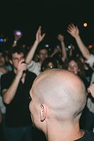 Nocturnal concert crowd cheering live music performance in euphoric ecstasy, young adults celebrating in nightlife club atmosphere, foreground shaved head of spectator observing ecstatic audience under dim stage illumination, bokeh lights dotting dark noc [IBR125446490]