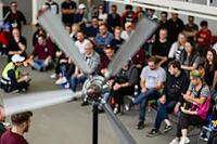 Engineering students attend aerodynamic wind turbine demonstration in technical university lecture hall, focus on mechanical rotor blades, blurred audience of young academics observing technological presentation, conceptual synergy between renewable energ [IBR125446493]