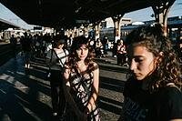 Urban isolation amidst crowded public transit, an introspective woman stands frozen on a sunlit train platform, deep contemplative vulnerability contrasted against moving commuter streams, rhythmic shadow patterns across concrete surfaces embody the psych [IBR125446495]
